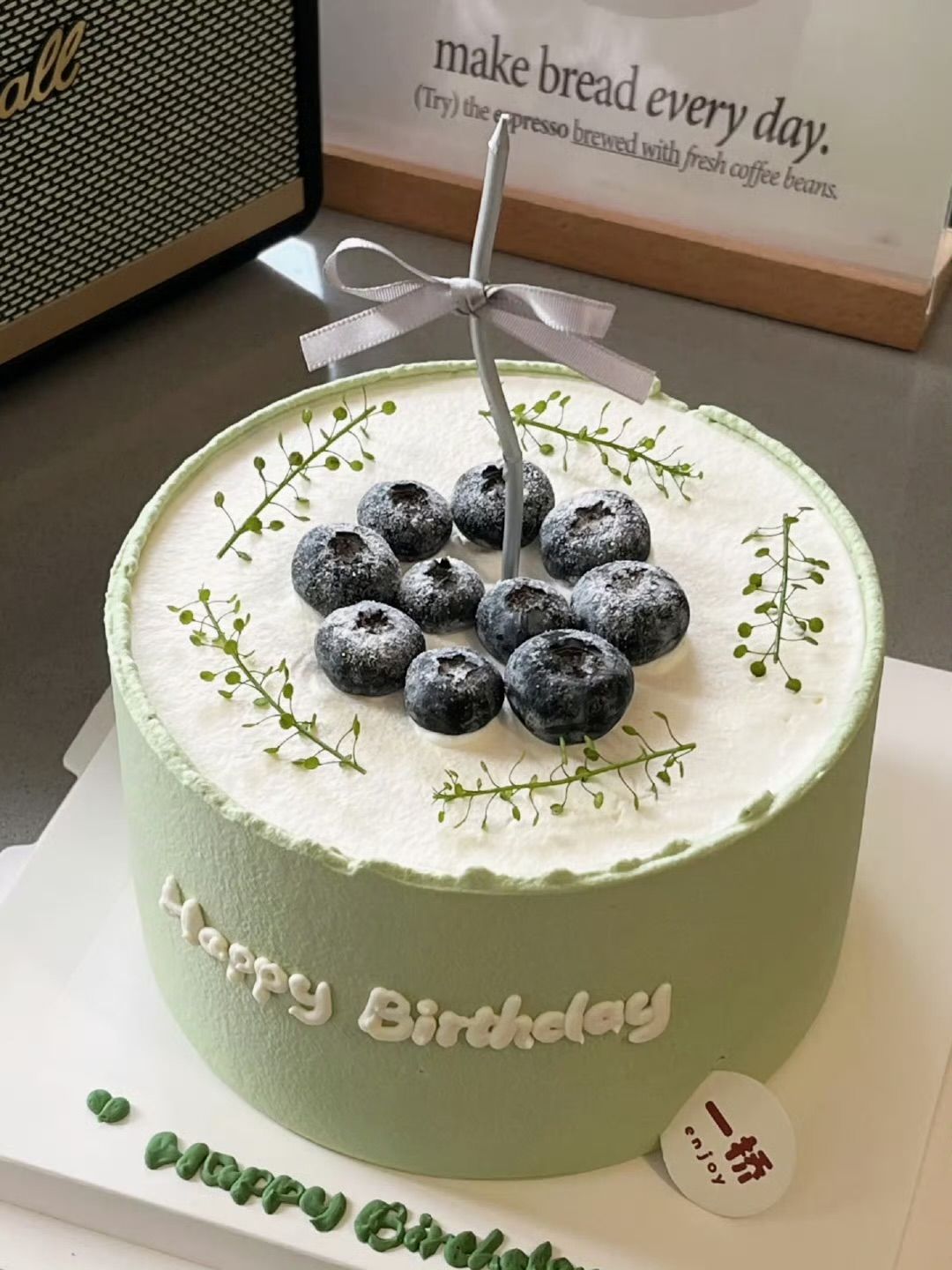 Minimalist round cake with sage green frosting on the sides, white cream on top, decorated with fresh blueberries, delicate green sprigs, and a silver candle with a grey ribbon.