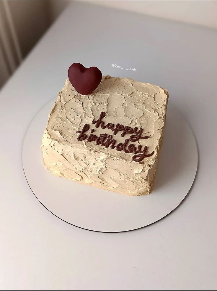A square-shaped birthday cake with thick, palette-knife textured beige frosting, featuring chocolate brown script and a single deep burgundy heart topper.
