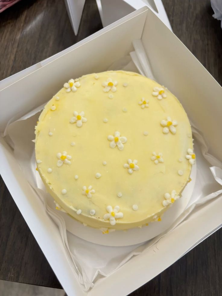 Top-down view of a round cake with pastel yellow frosting and small white buttercream daisies in a white bakery box.