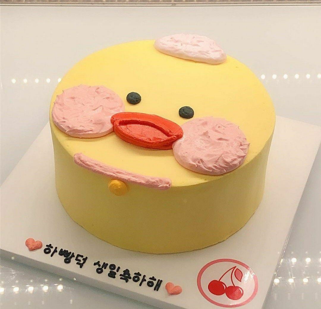 A round birthday cake decorated to look like a yellow duck with pink frosted cheeks, an orange beak, and Korean text on the cake board.