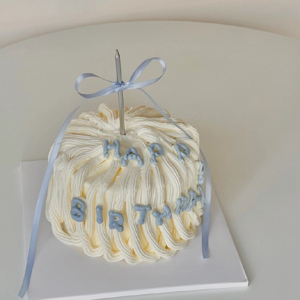 A round cake covered in heavy vertical white buttercream ridges, topped with a single silver candle tied with a light blue ribbon and "Happy Birthday" in blue bubble lettering.