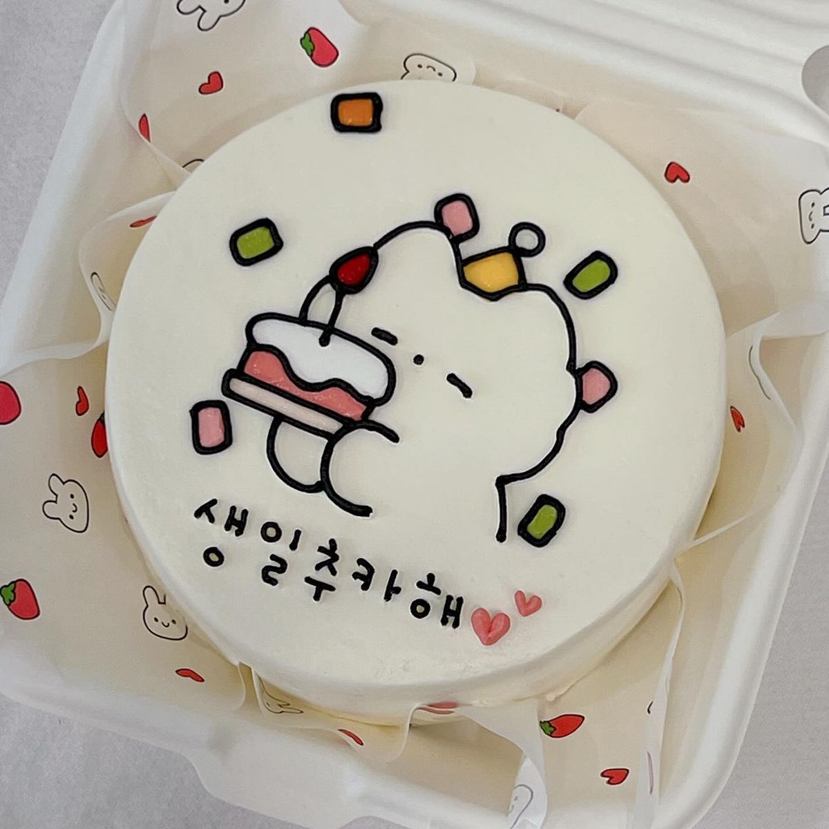 Cute white Korean bento cake featuring a hand-drawn bear character holding a small cake slice with colorful confetti and Korean birthday text.