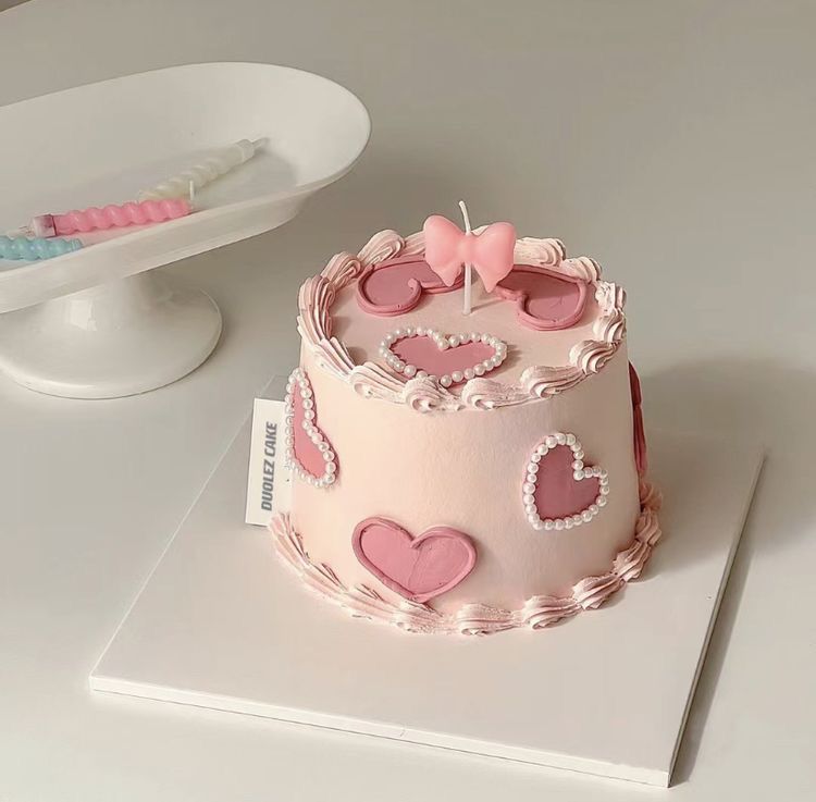 Aesthetic light pink round cake decorated with darker pink frosting hearts, edible pearls, and a pink bow candle on a white surface.
