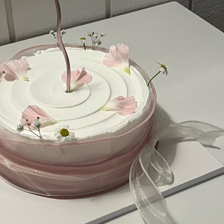 Close-up of a white frosted cake wrapped in sheer pink and white ribbons, topped with delicate pink flower petals and tiny white daisies.