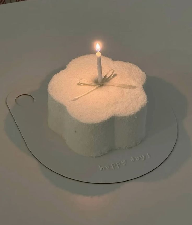 A minimalist flower-shaped white cake with a fuzzy textured frosting, a single lit candle tied with a delicate cream bow, and "happy day!" written on the cake board.