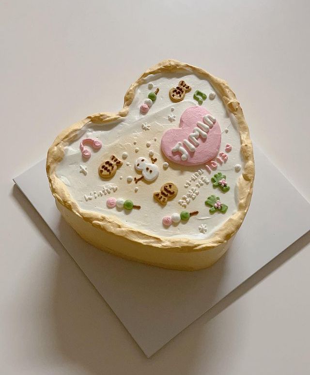 A heart-shaped cake with a rustic beige border, white frosting top decorated with a snowman, fish-shaped pastries, dango skewers, and a pink heart containing the name Jimin.