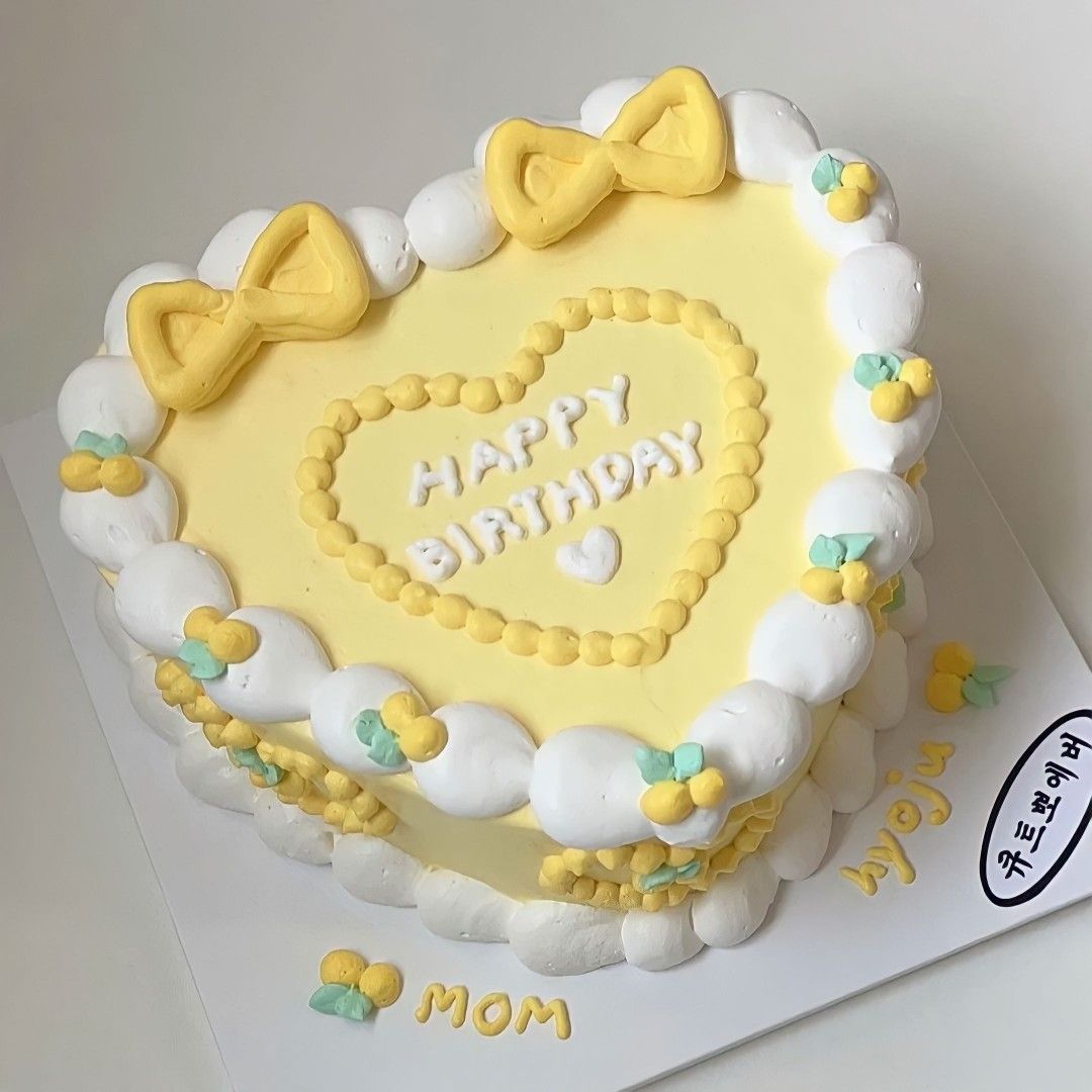 Heart-shaped birthday cake with pale yellow frosting, white cream dollops, yellow fondant bows, and "Happy Birthday" written in the center.