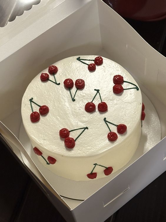 Minimalist round white buttercream cake decorated with a repetitive pattern of small red piped cherries with green stems in a white bakery box.