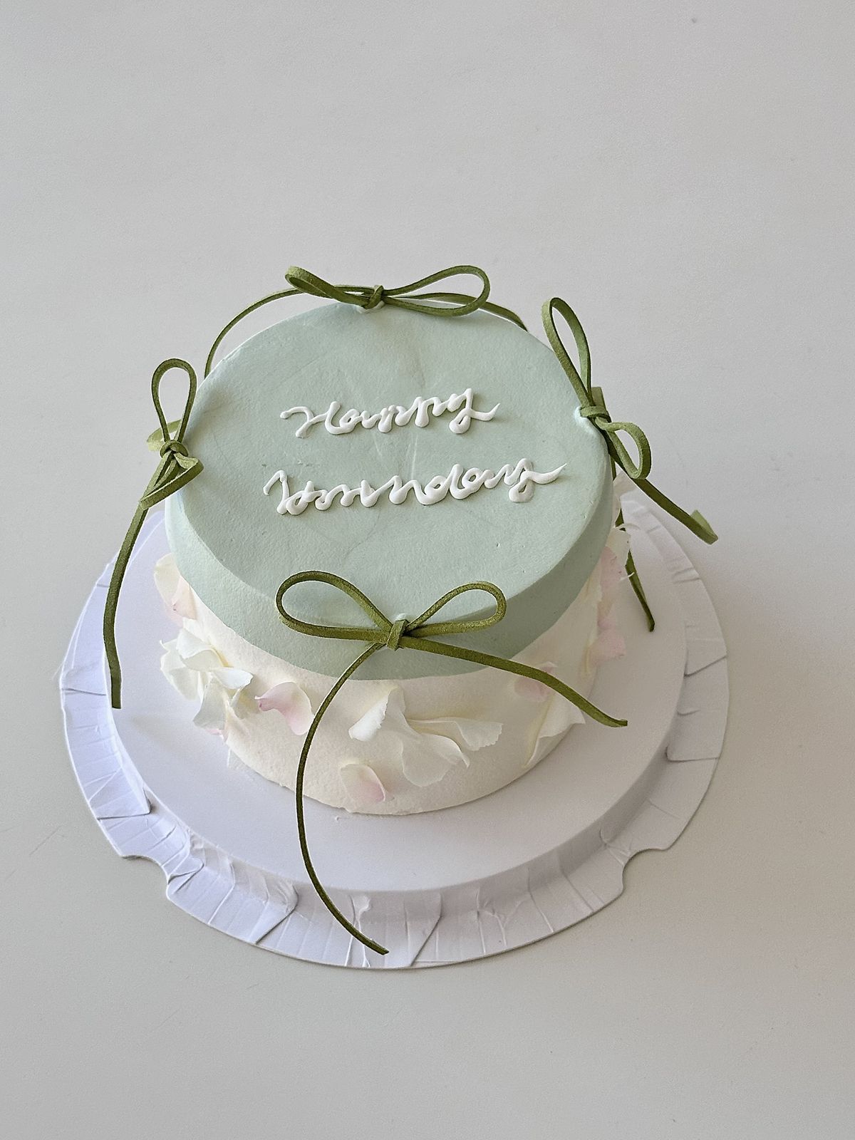 Minimalist two-tone cake with a mint green top and white base, featuring "Happy Birthday" script, delicate white flower petals, and slender olive green ribbon bows.