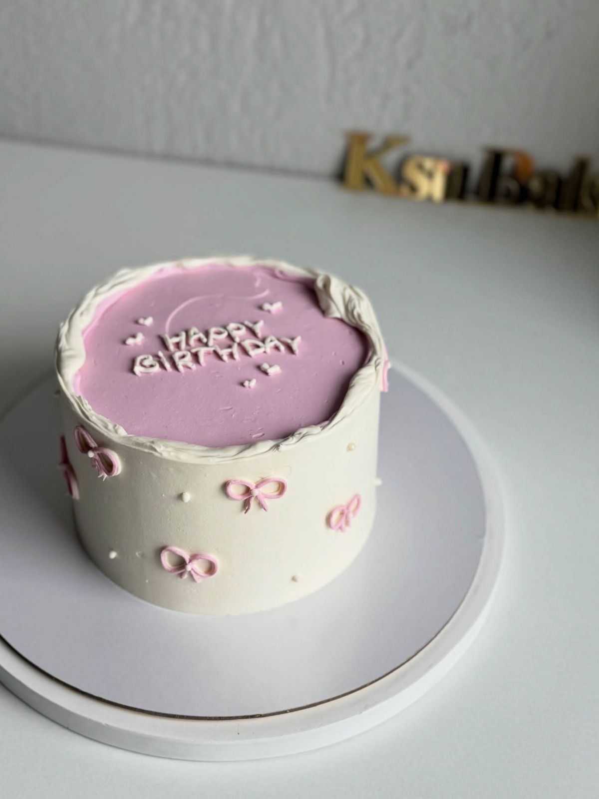A round white frosted cake with a lavender purple top, featuring "Happy Birthday" in white script and dainty pink piped bows on the sides.