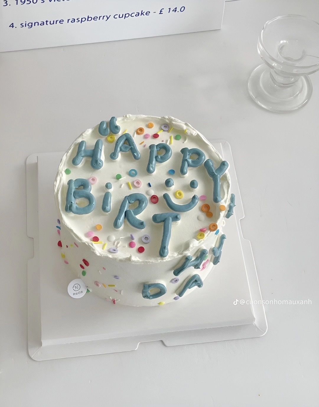 A round white frosted cake topped with multi-colored confetti sprinkles and "Happy Birthday" written in thick, 3D blue-grey buttercream bubble letters.