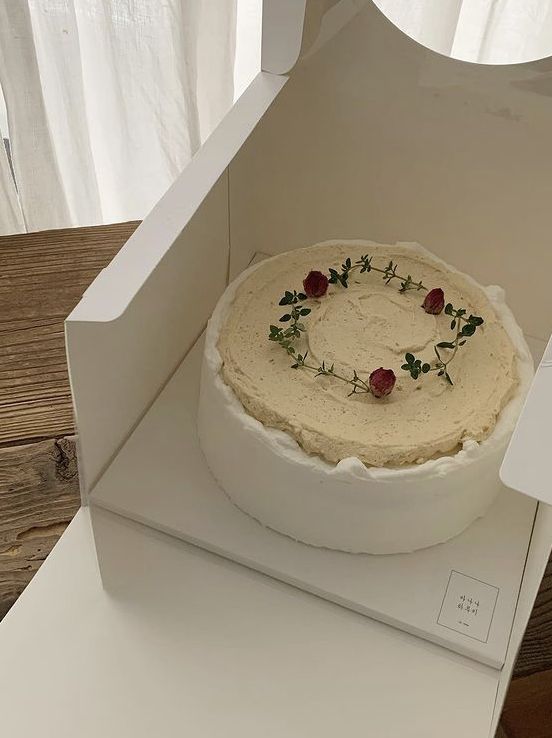 A round cake with smooth white sides and a textured beige top, decorated with a circular wreath made of fresh green thyme sprigs and tiny dried red rosebuds in a white bakery box.