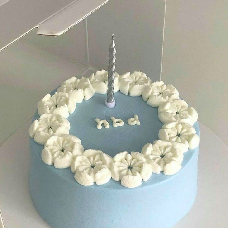 A round cake with smooth pastel blue frosting, decorated with a wreath of white buttercream flowers and a single silver candle in the center.