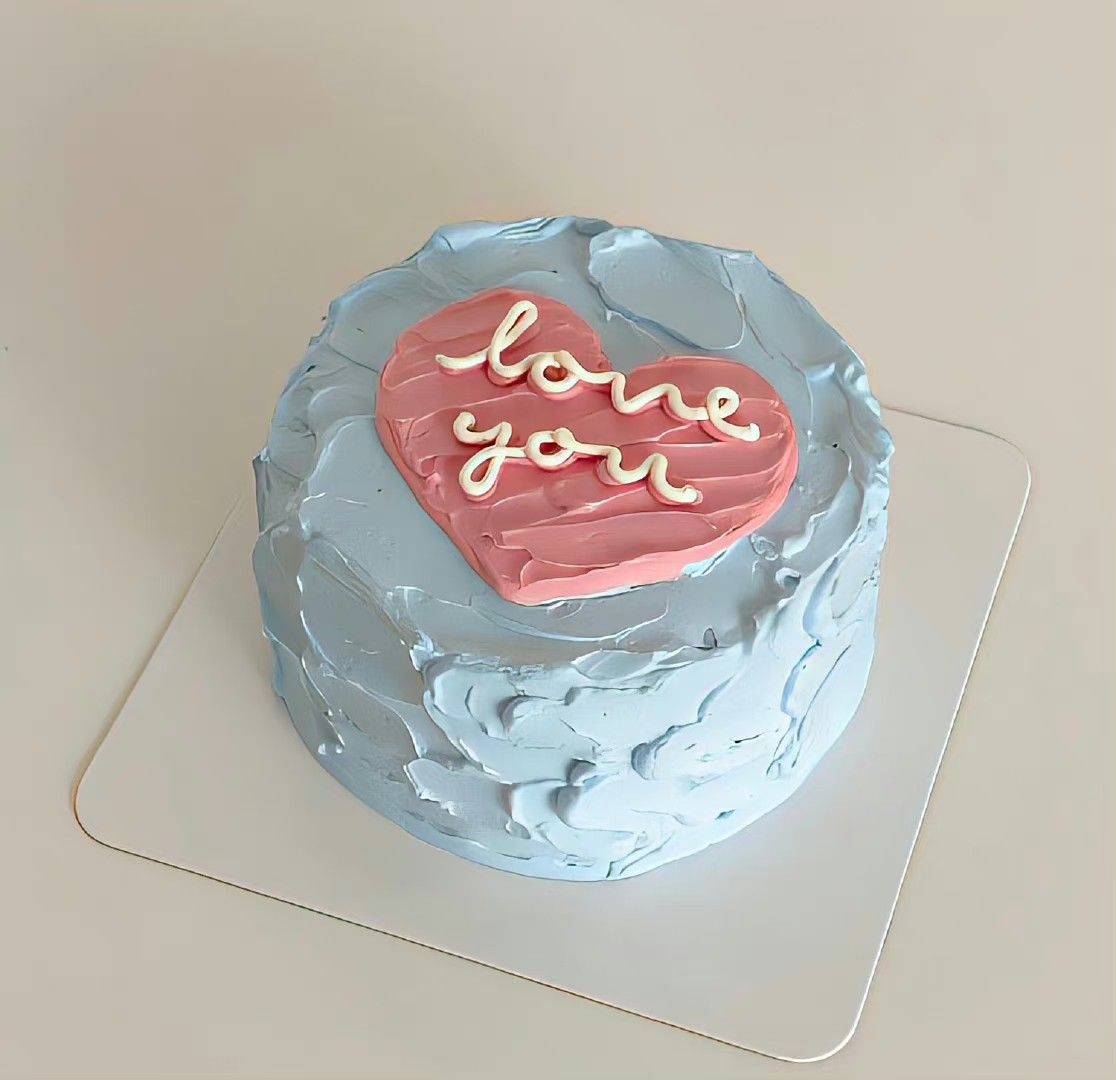 Rustic textured light blue bento cake with a pink buttercream heart and white cursive 'love you' icing on a white square board.