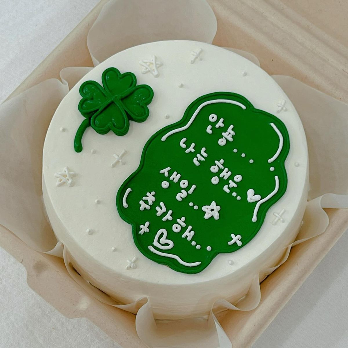 Minimalist white Korean bento cake featuring a green four-leaf clover illustration and hand-piped Korean birthday message in a bento box.