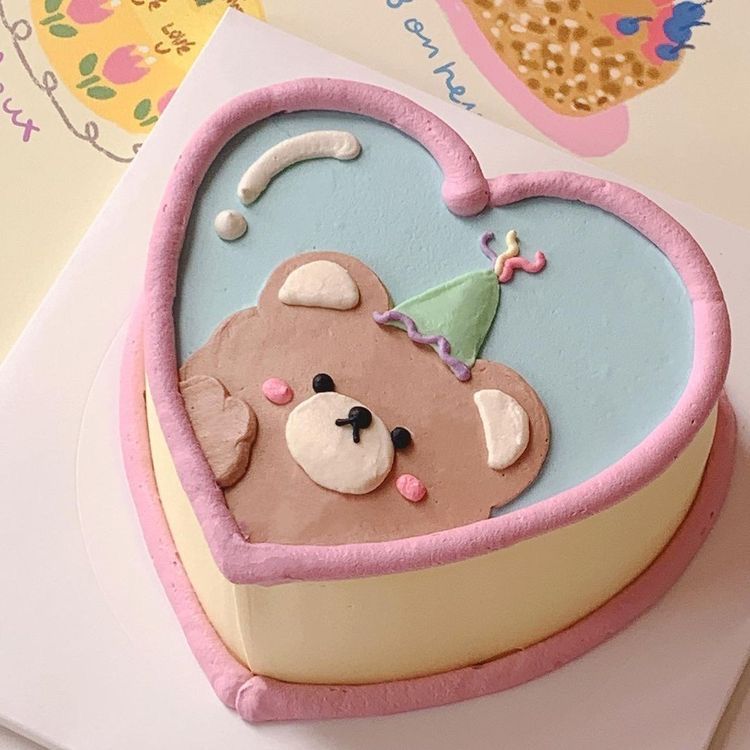 Heart-shaped pastel cake with a brown bear wearing a party hat, light blue frosting, and pink borders for aesthetic birthday inspiration.