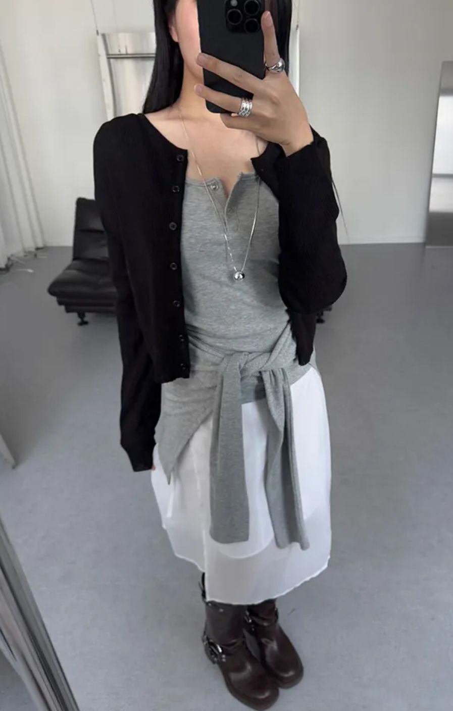 A woman takes a mirror selfie wearing an open black cardigan, a grey longline henley top tied at the waist, a white sheer midi skirt, and dark brown leather biker boots with buckle details.