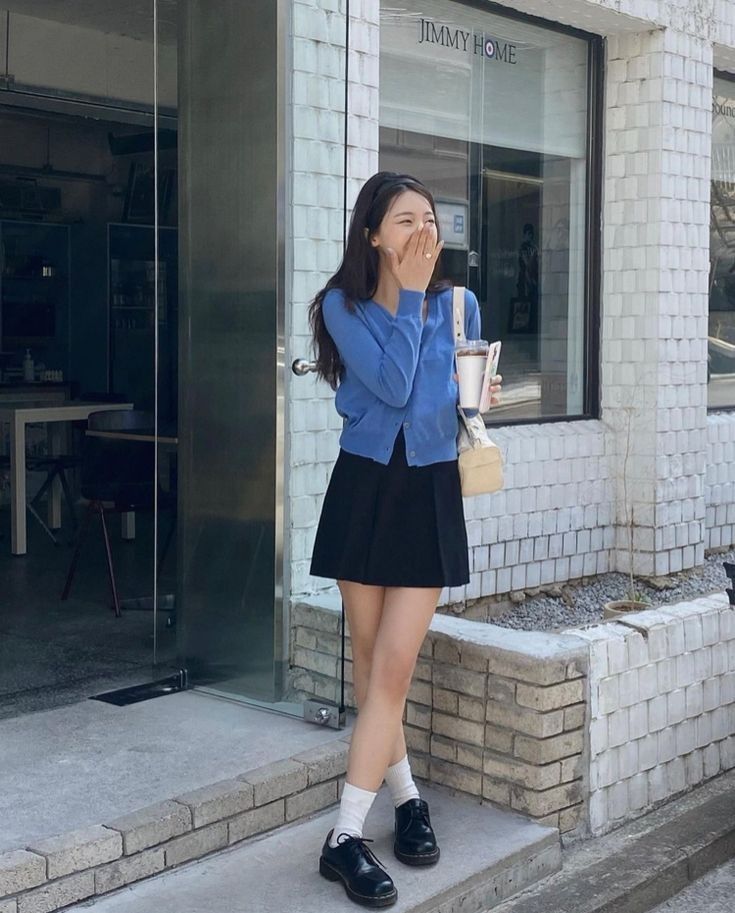 A woman laughing outside a white brick building wearing a buttoned blue cardigan, a black pleated mini skirt, white socks, and black leather oxford shoes while carrying a cream bag.