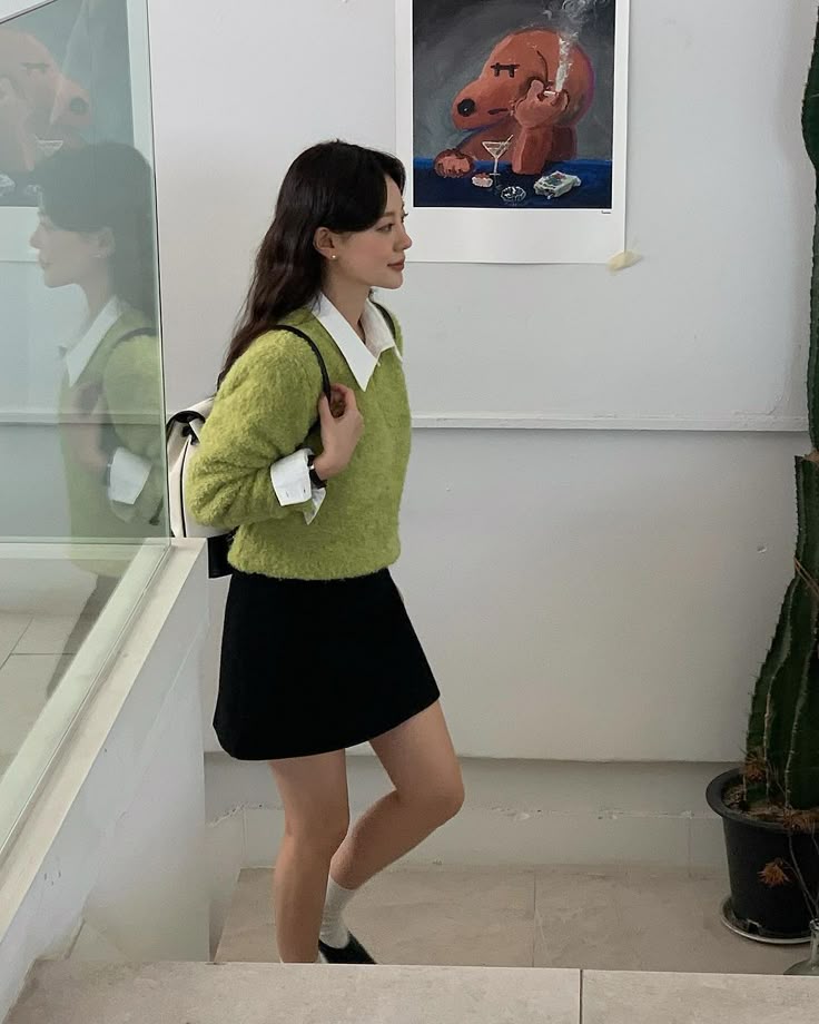 A woman walks through a white-walled gallery wearing a fuzzy lime green v-neck sweater over a white button-down shirt, a black mini skirt, and a white backpack.
