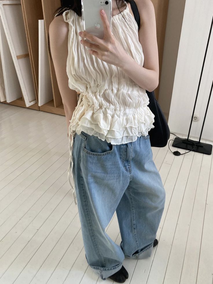 A woman takes a mirror selfie in a studio wearing a cream ruffled halter-style top with smocking details, oversized light blue denim jeans, and a black shoulder bag.