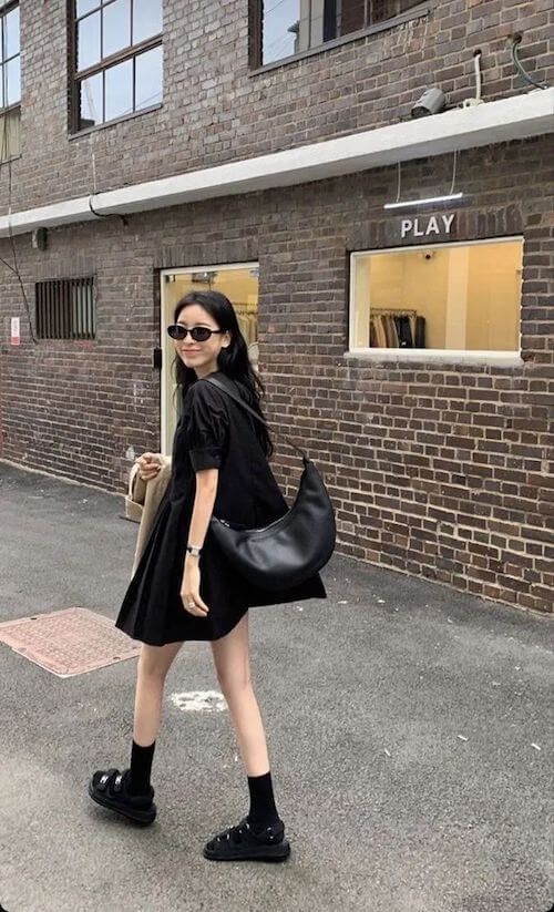 A woman walking in a city wearing a black puff-sleeve mini dress, black crew socks, chunky sandals, and a large black crescent crossbody bag.
