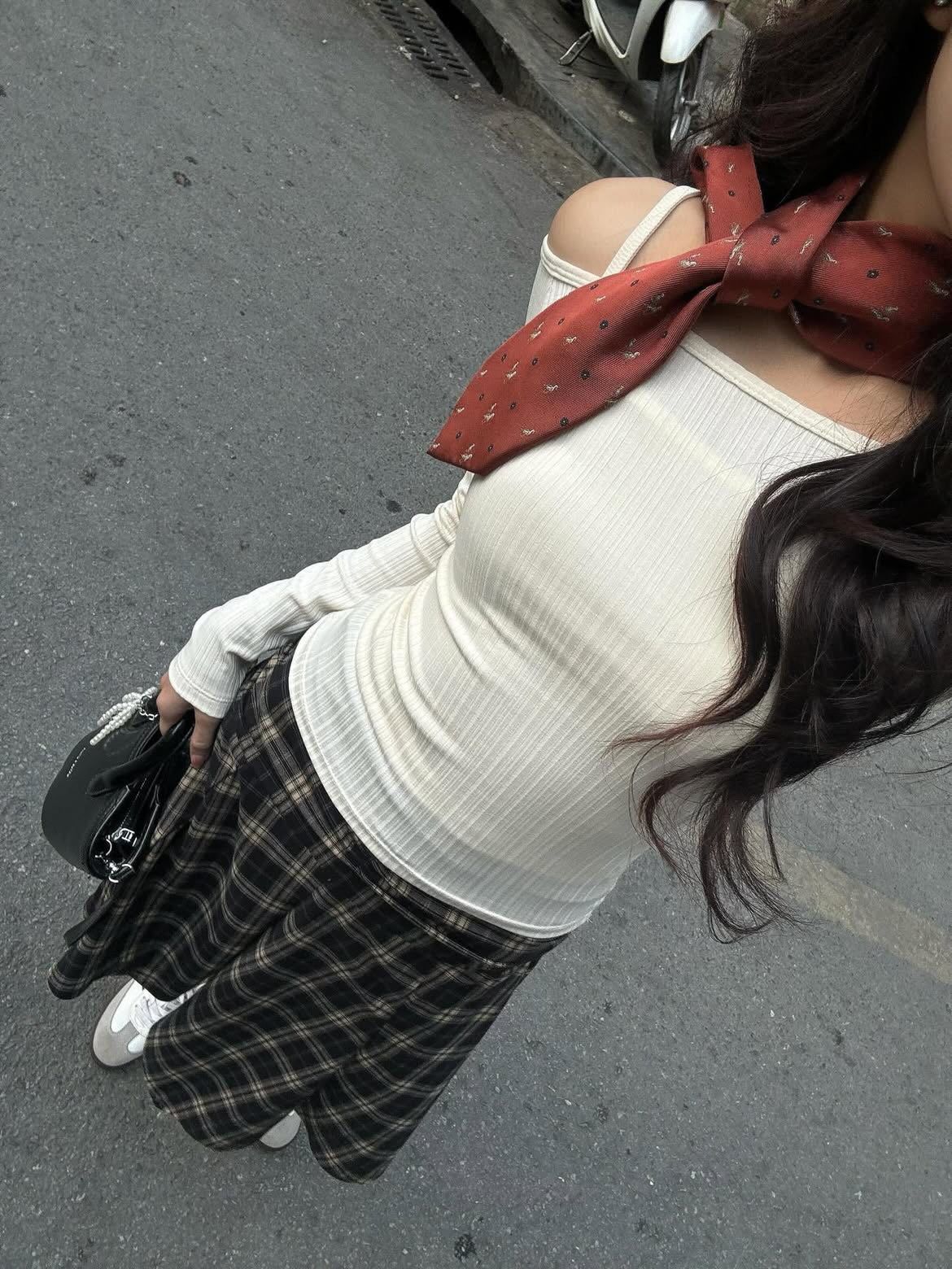 A woman stands on a street wearing a long-sleeved cream ribbed top with an off-the-shoulder detail, a dark plaid mini skirt, a rust-colored patterned neck scarf, and white sneakers while carrying a black bag.