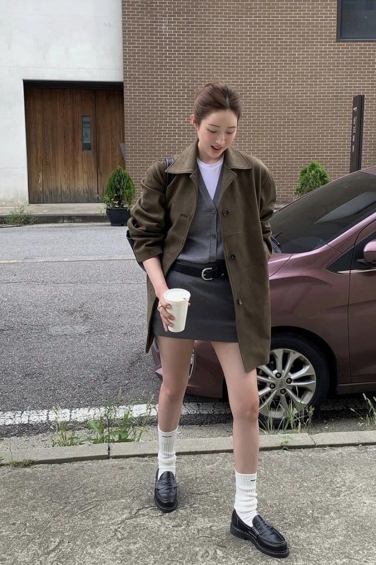 A woman walking outside holding a coffee cup, wearing an olive green jacket over a grey cardigan, a dark grey mini skirt with a black belt, white slouchy socks, and black loafers.