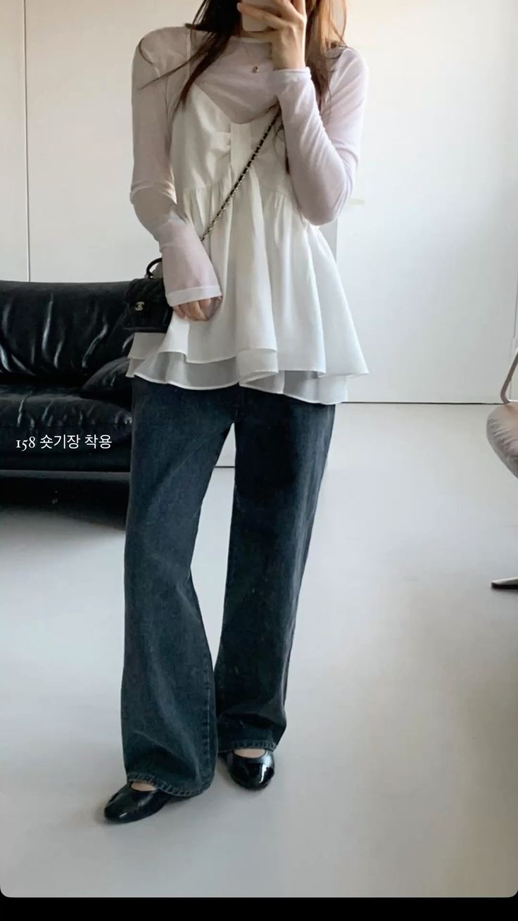 A woman stands in a minimalist room wearing a white tiered ruffle camisole over a sheer white long-sleeve top, dark wide-leg jeans, and black ballet flats while carrying a small black quilted chain bag.
