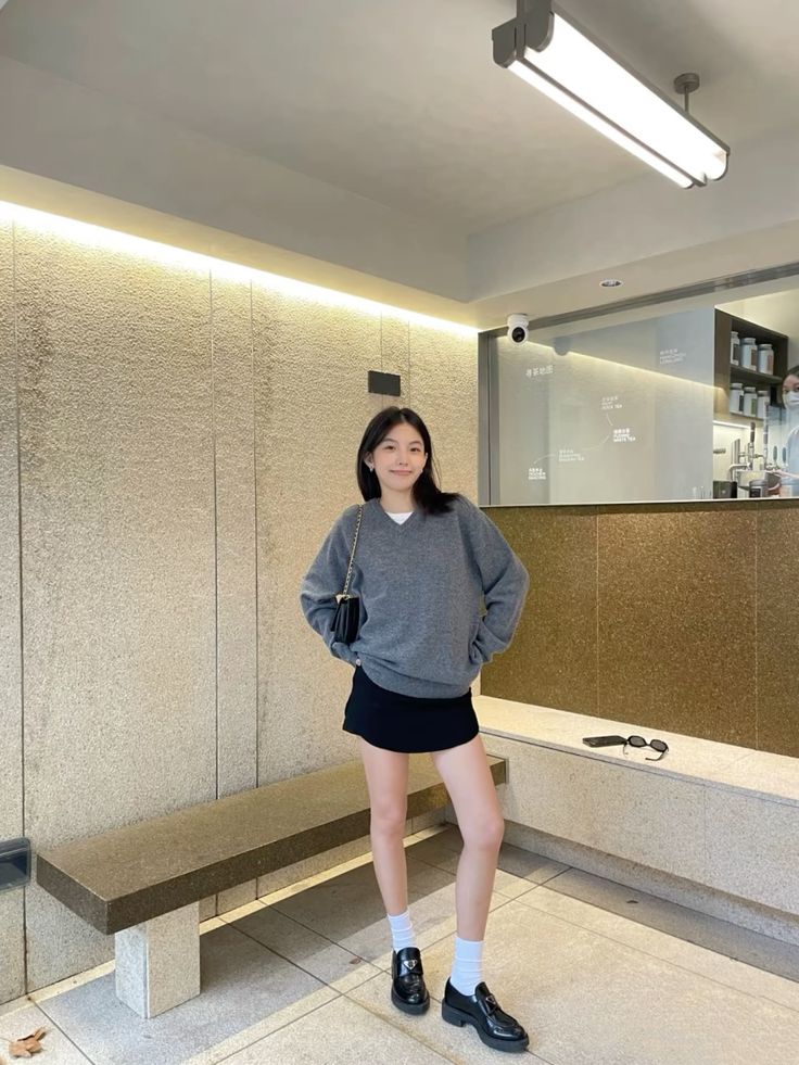 A woman in a cafe wearing an oversized grey v-neck sweater, a short black skirt, white crew socks, and chunky black loafers with a small black crossbody bag.
