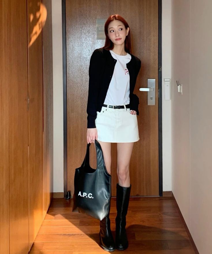 A woman stands indoors wearing a black cardigan over a white t-shirt, a white denim mini skirt with a black belt, knee-high black leather boots, and carrying a black A.P.C. tote bag.