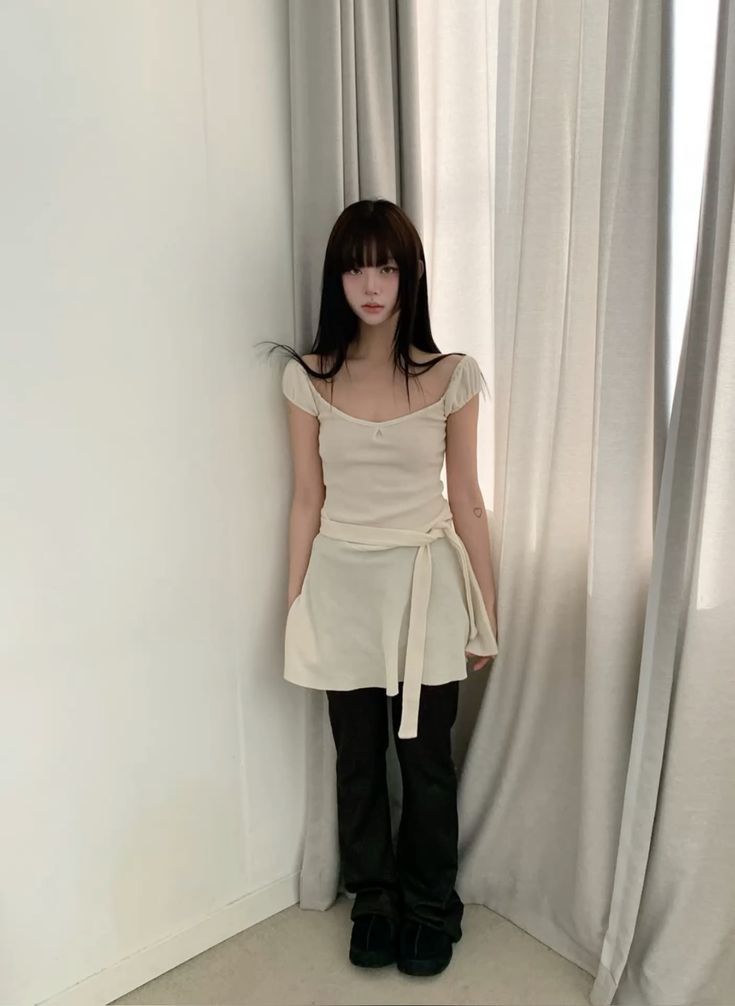 A woman with long dark hair and bangs stands against a white wall wearing a cream-colored cap-sleeve tunic with a waist tie and black flared pants.