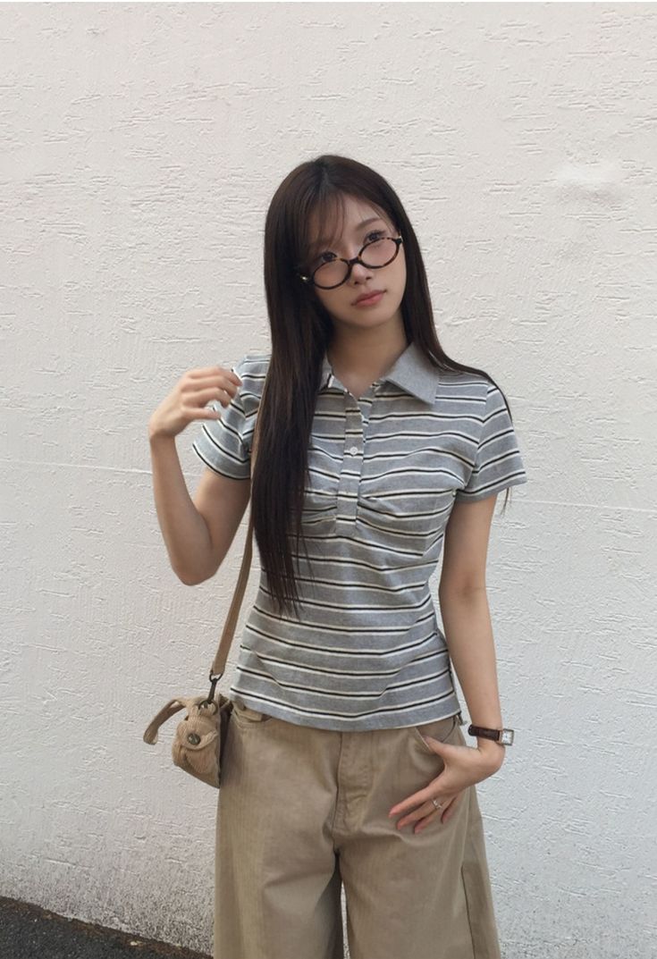 A woman with long dark hair wearing tortoiseshell glasses, a grey and white horizontal striped polo shirt, and tan wide-leg khaki trousers with a small beige crossbody bag.