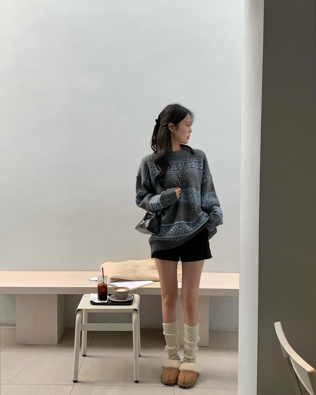 A woman in a minimalist cafe wearing an oversized grey patterned sweater, black shorts, cream slouchy leg warmers, and tan shearling slippers.