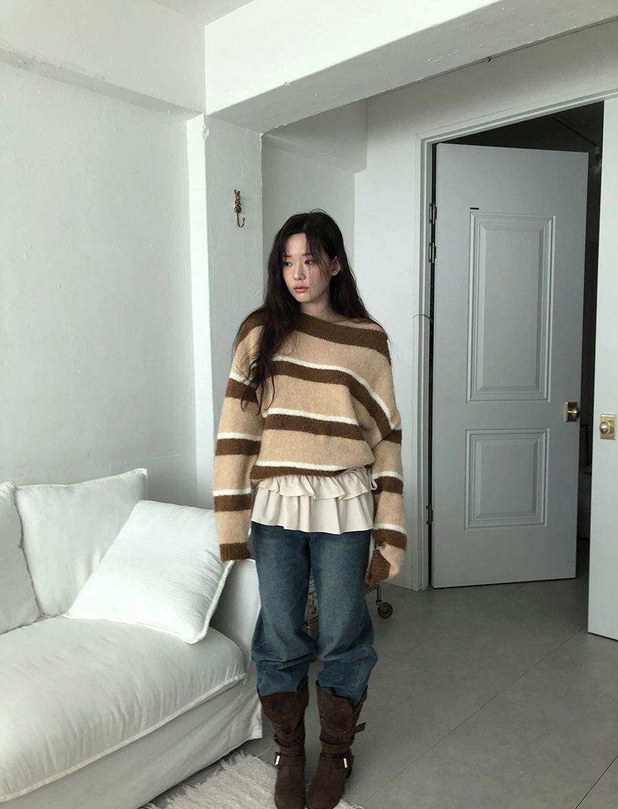 A woman in a white room wearing an oversized tan and brown striped sweater with a ruffle trim, baggy blue jeans, and tall brown suede boots.