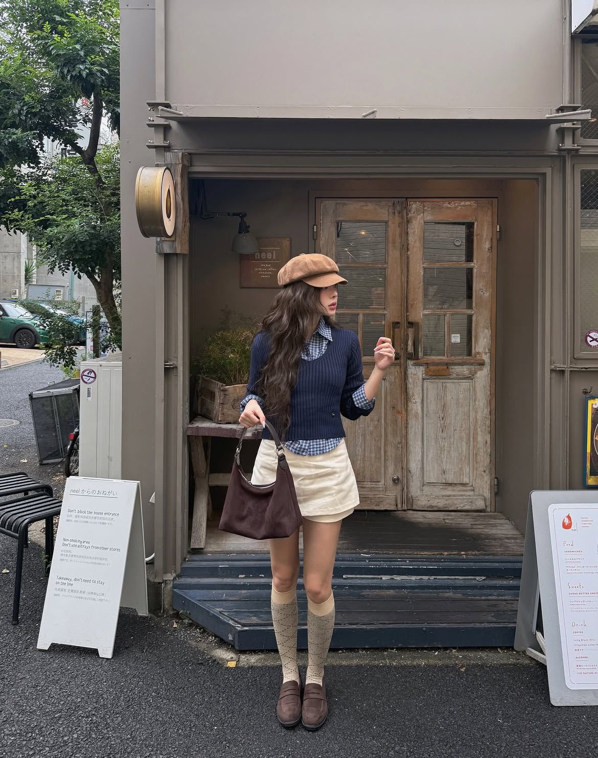 A woman stands in front of a rustic cafe wearing a navy blue ribbed sweater vest over a plaid shirt, a cream mini skirt, knee-high socks, brown loafers, and a tan newsboy cap.