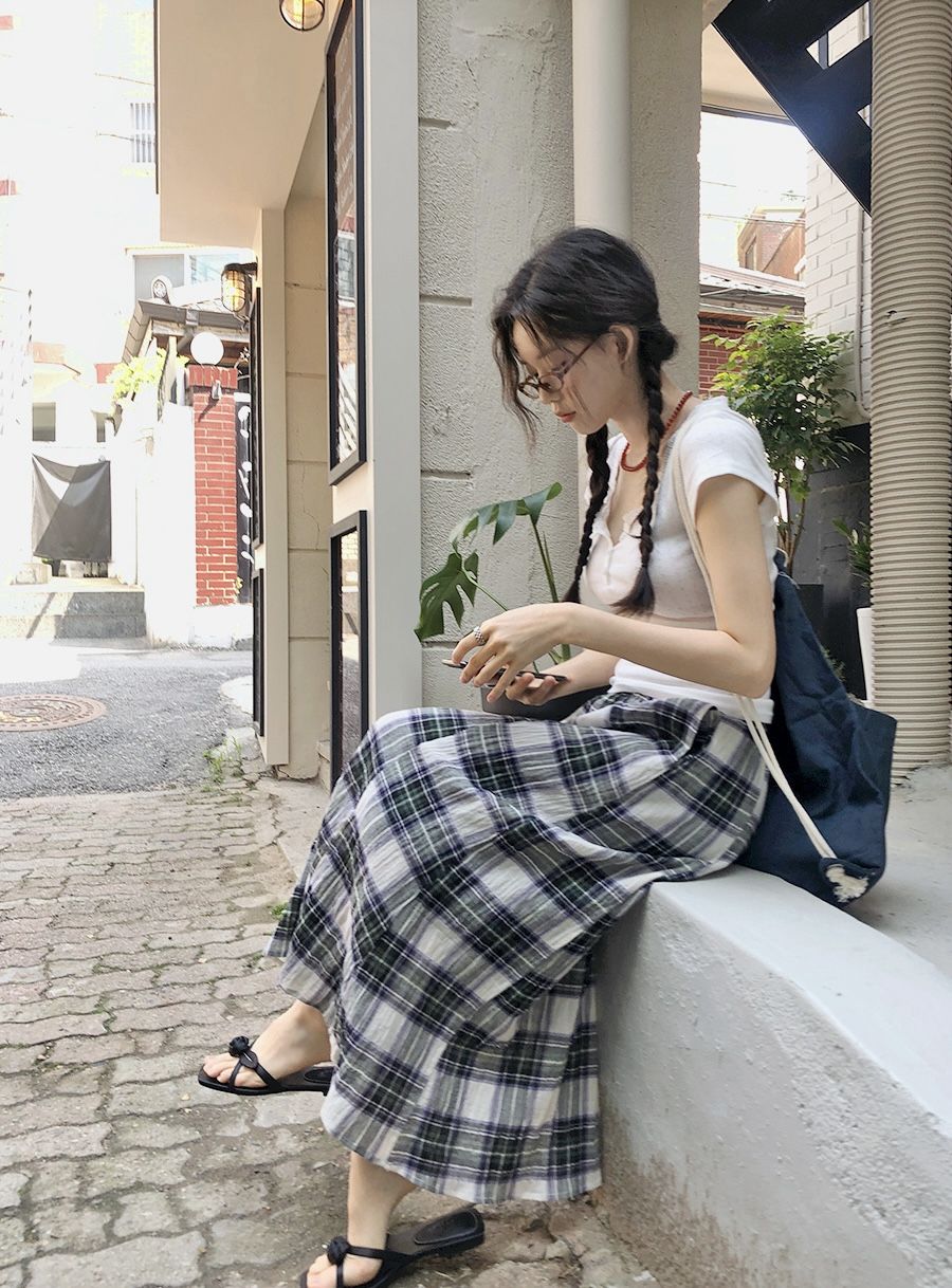A woman with braided hair sitting outdoors wearing a white short-sleeved henley t-shirt and a long navy and green plaid maxi skirt with black sandals.