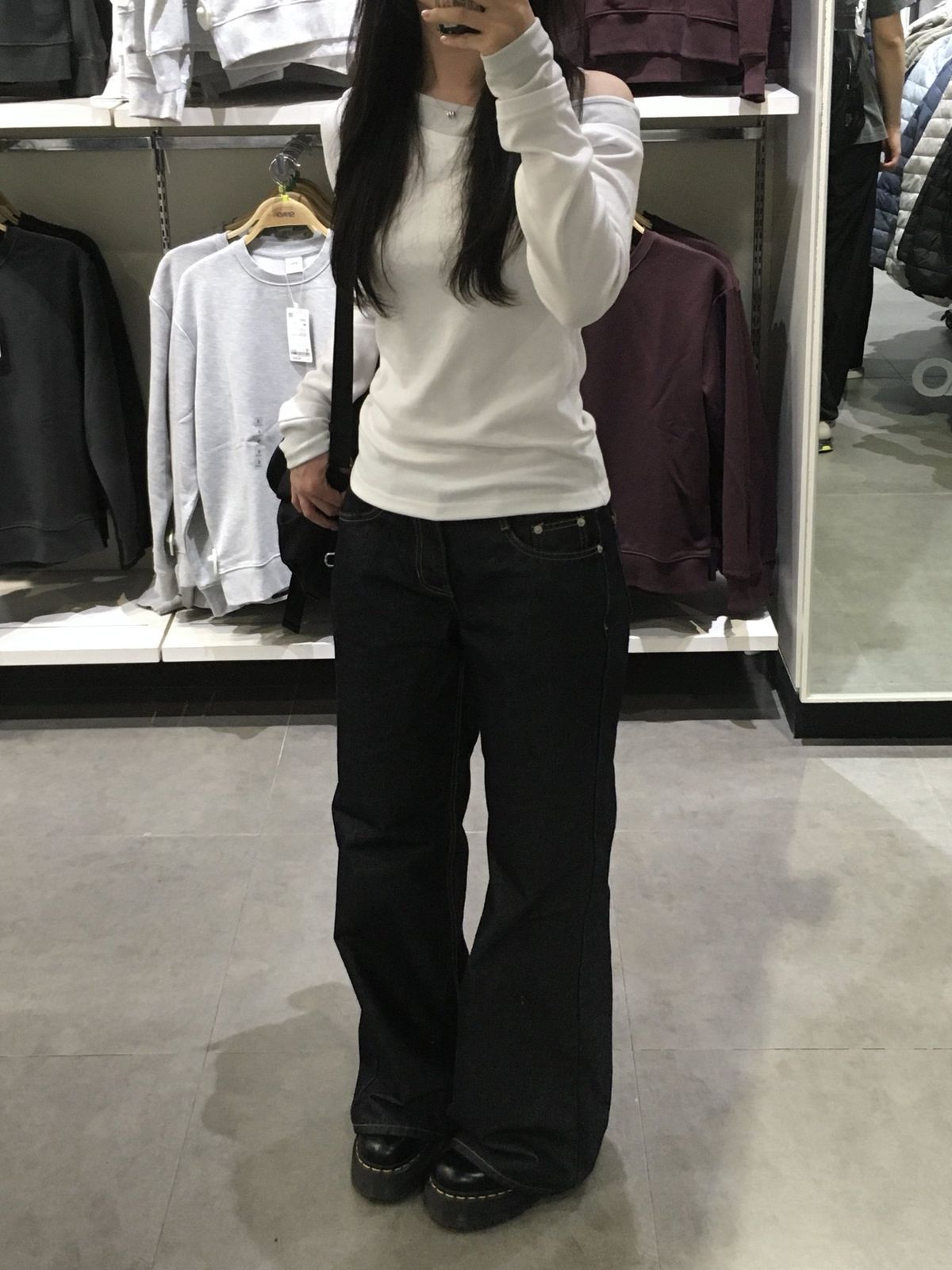A woman in a clothing store wearing a white off-the-shoulder long-sleeve top over a grey layer paired with wide-leg dark denim jeans and black platform boots.