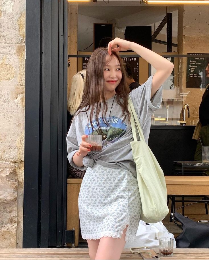 A woman smiling at an outdoor cafe wearing an oversized grey graphic t-shirt knotted at the waist, a light blue floral mini skirt with lace trim, and a lime green tote bag.