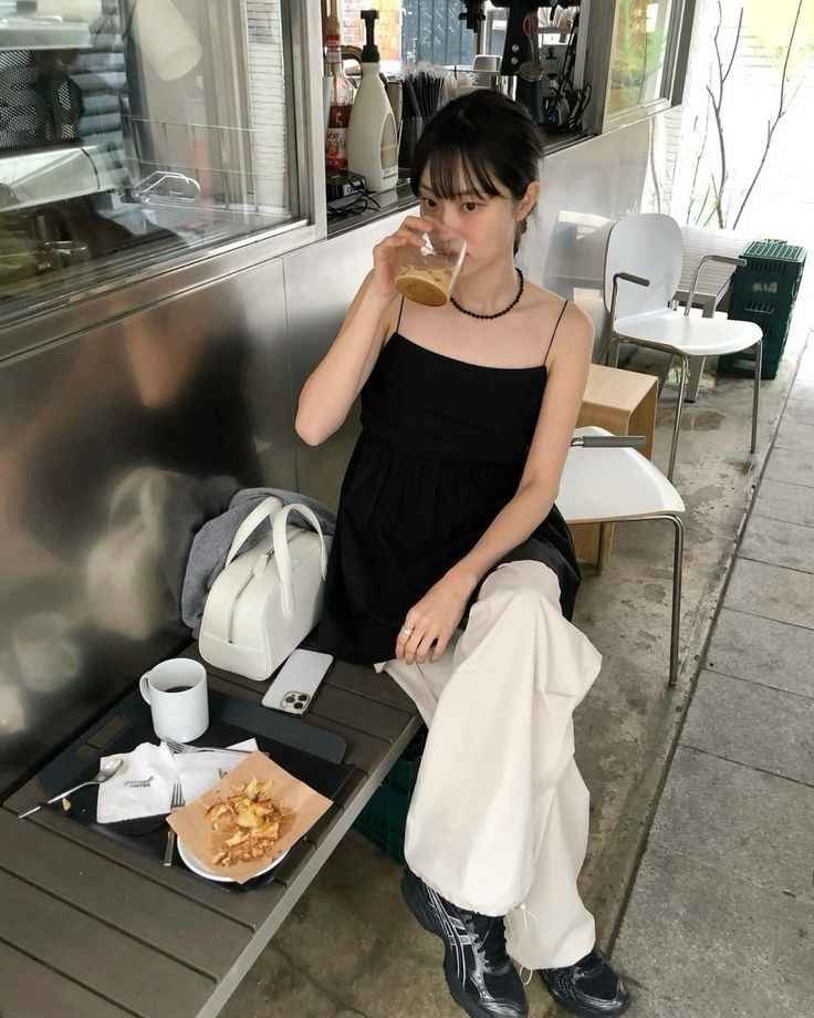 A woman sitting at an outdoor cafe drinking coffee, wearing a black babydoll camisole, white wide-leg trousers, black sneakers, and carrying a white leather bag.