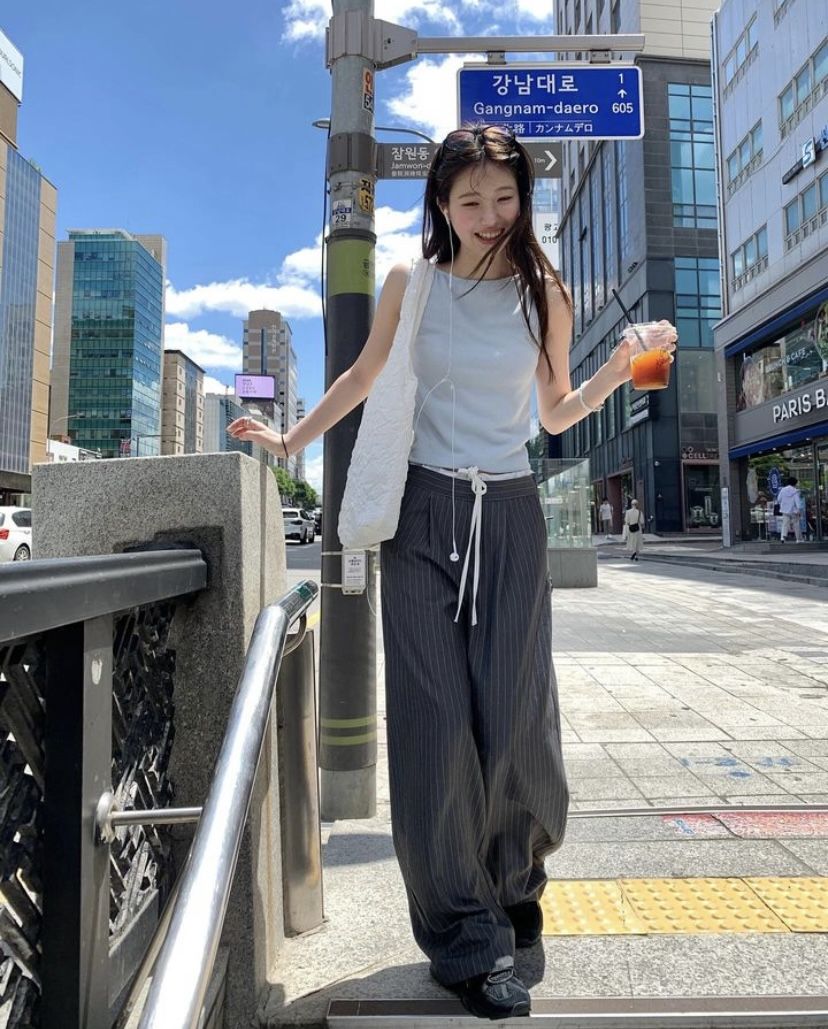 A woman walking in a city wearing a light blue sleeveless tank top, dark grey pinstripe wide-leg trousers with a drawstring, and a white quilted bag.