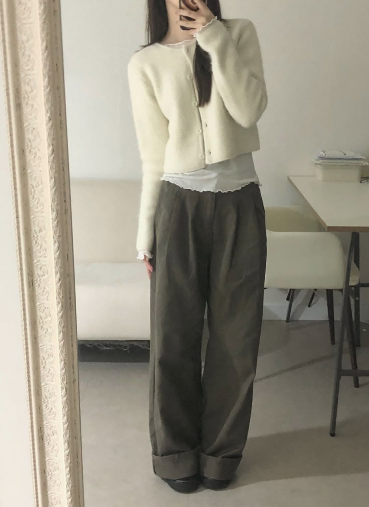 A woman in a mirror selfie wearing a fluffy cream button-up cardigan over a white ruffled top with oversized charcoal grey pleated trousers.