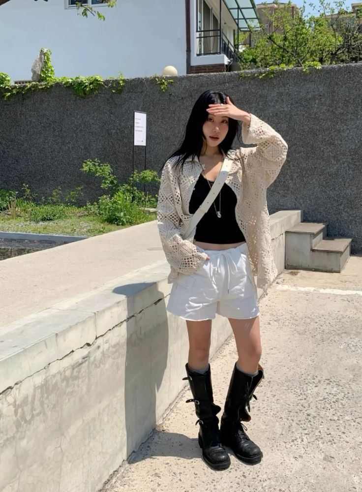 A woman standing outdoors wearing an oversized cream crochet button-down shirt over a black tank top and white drawstring shorts, styled with tall black buckled boots and a white crossbody bag.