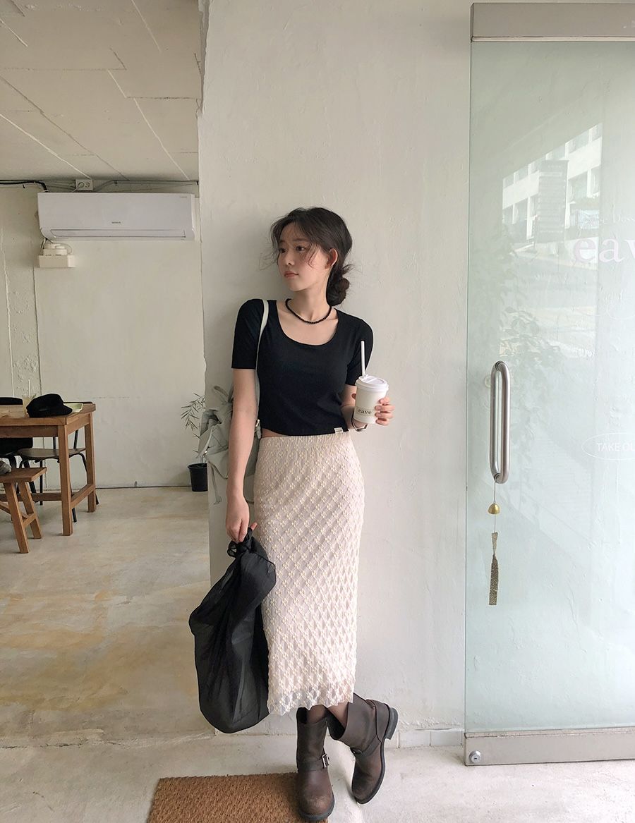 A woman standing in a cafe wearing a black short-sleeved crop top, a cream lace column midi skirt, and distressed brown leather biker boots.