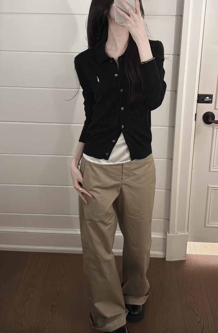 A woman posing in a mirror wearing a black button-down cardigan over a cream top with baggy khaki trousers and black platform shoes.