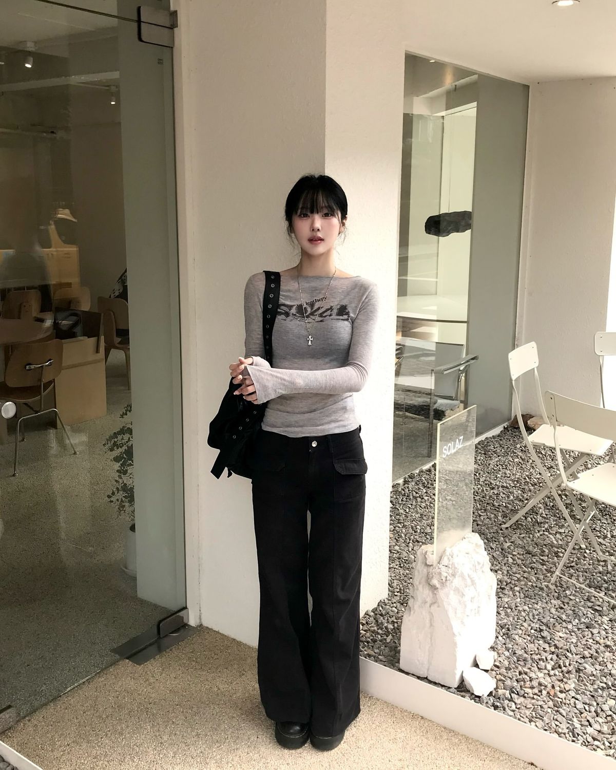 A woman standing in a modern cafe wearing a grey graphic long-sleeve boatneck top with black flare cargo trousers and a studded black bag.