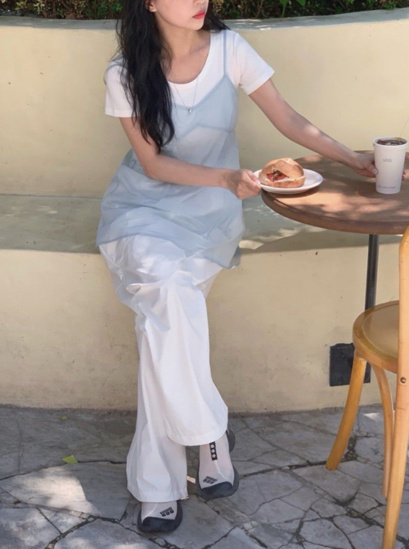 A woman sitting at a cafe table wearing a sheer light blue camisole over a white t-shirt, paired with white wide-leg trousers and white sneakers with black accents.