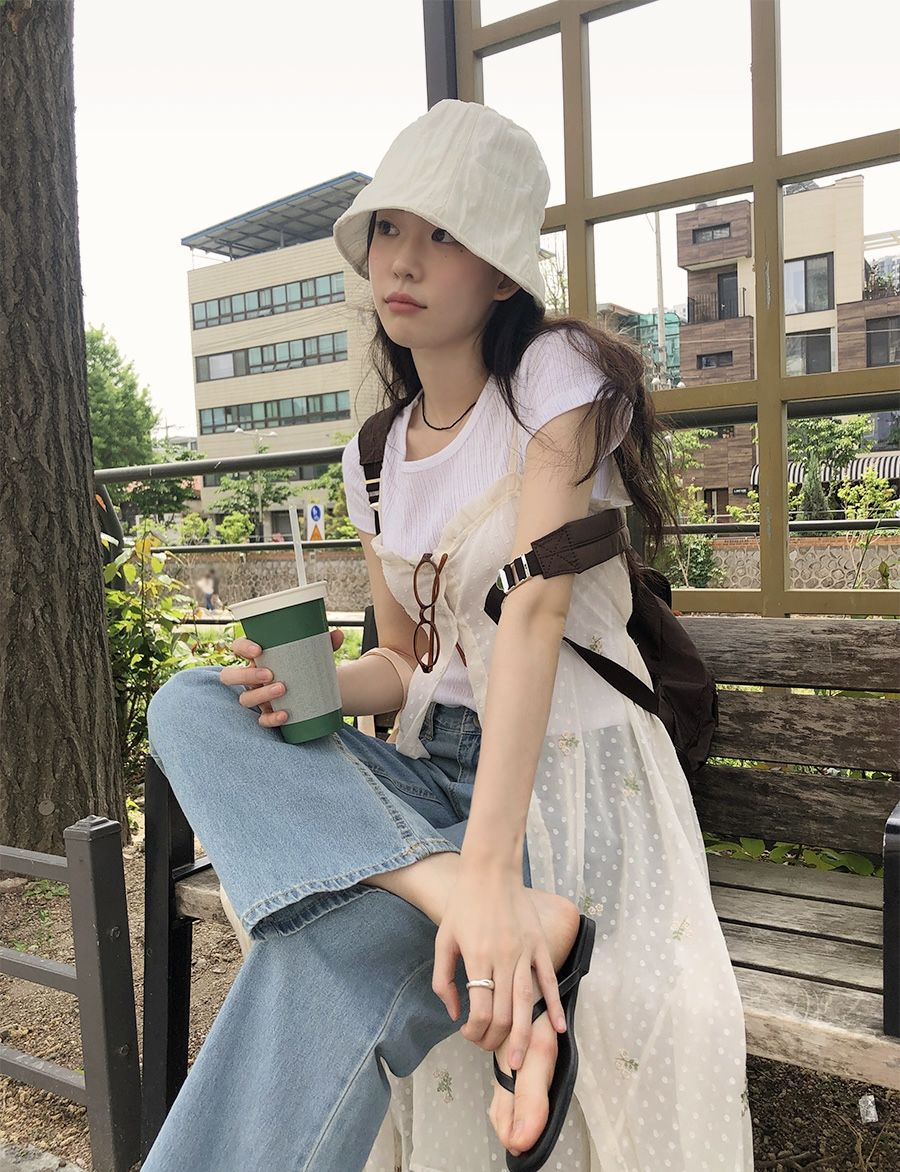 A woman sitting on a park bench wearing a sheer white polka dot tunic over a white t-shirt and wide-leg blue jeans, styled with a white bucket hat and brown backpack.