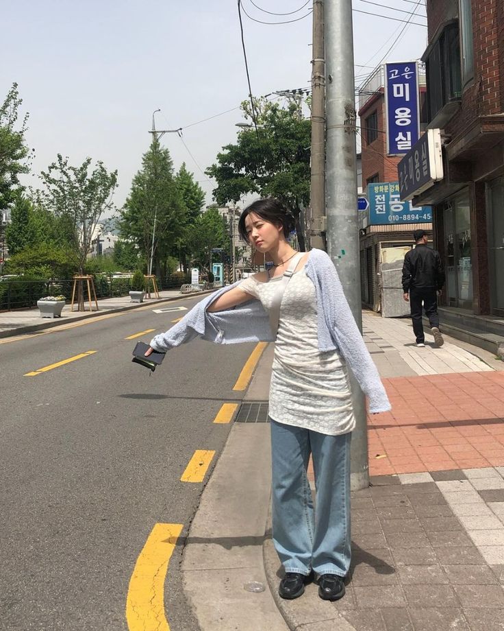 A woman standing on a city street wearing a cream floral lace mini dress over baggy blue jeans, paired with a light purple shrug and black loafers.