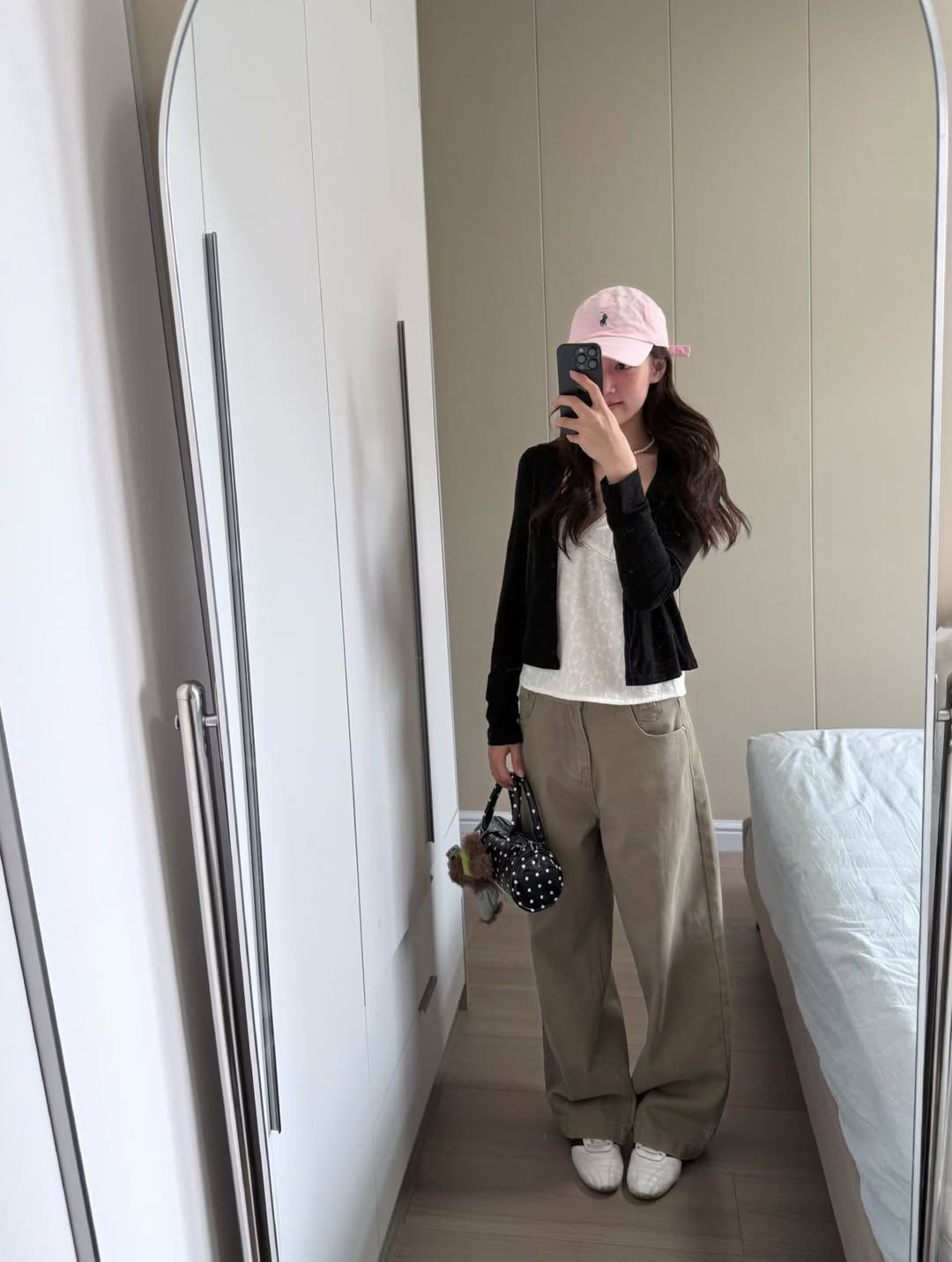 A student in a mirror selfie wearing a white lace camisole, a black cropped cardigan, wide-leg khaki pants, and a pink baseball cap.