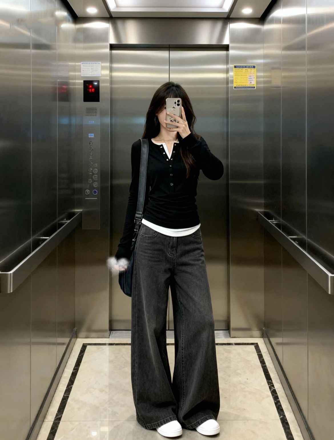 A student taking an elevator mirror selfie wearing a black button-up henley with a white layer underneath, wide-leg dark grey jeans, and white sneakers.